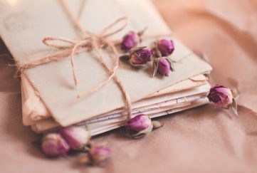 Best Love Letters Of All Time. Images features a stack of enveloped and folded love letters tied in a string bow and sprinkled with tiny pink rose buds.