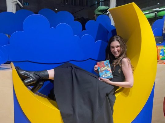 Katherine Rundell attending a book signing at Piccadilly Waterstones. She is sitting on a large half moon set piece and holding her children's novel, Impossible creatures. She smiling wide and wearing a long black dress and black boots.