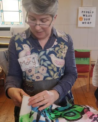 Mary Morton, founder of the Edinburgh Street Stitchers. Mary is seen sitting in a community hall stitching a repair while wearing a dress covered in little patchworks.