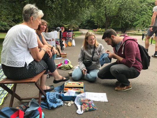 Camping chairs are lined along a park path with street stitchers. A young couple have sat down on the path to join in and are stitching repairs of their own.