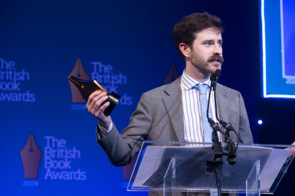 G.T. Karber receiving one of three prizes at The British Book Awards 2024. he's wearing a grey suit with a strop shirt and pale blue tie. He's making a speech while holding his award.