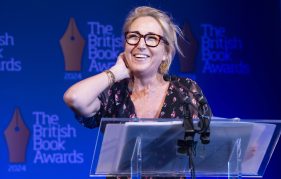 Lisa Jewell at The British Book Awards 2024 collecting one of her two awards. She is standing at the podium and smiling wide with her hand nervously on her neck. She's wearing her hair tied back, black framed glasses and a floral blouse.