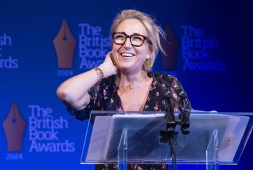 Lisa Jewell at The British Book Awards 2024 collecting one of her two awards. She is standing at the podium and smiling wide with her hand nervously on her neck. She's wearing her hair tied back, black framed glasses and a floral blouse.
