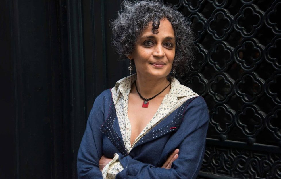 Arundhati Roy standing with her arms crossed wearing a blue cardigan and smiling.