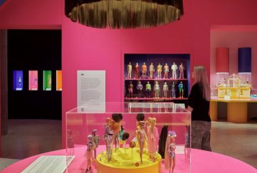 Photo from inside the Barbie exhibition London. There is a chandelier made of barbie hair, every wall is Barbie pink, and there are dolls of all variety in different types of casing spaced out across the room.