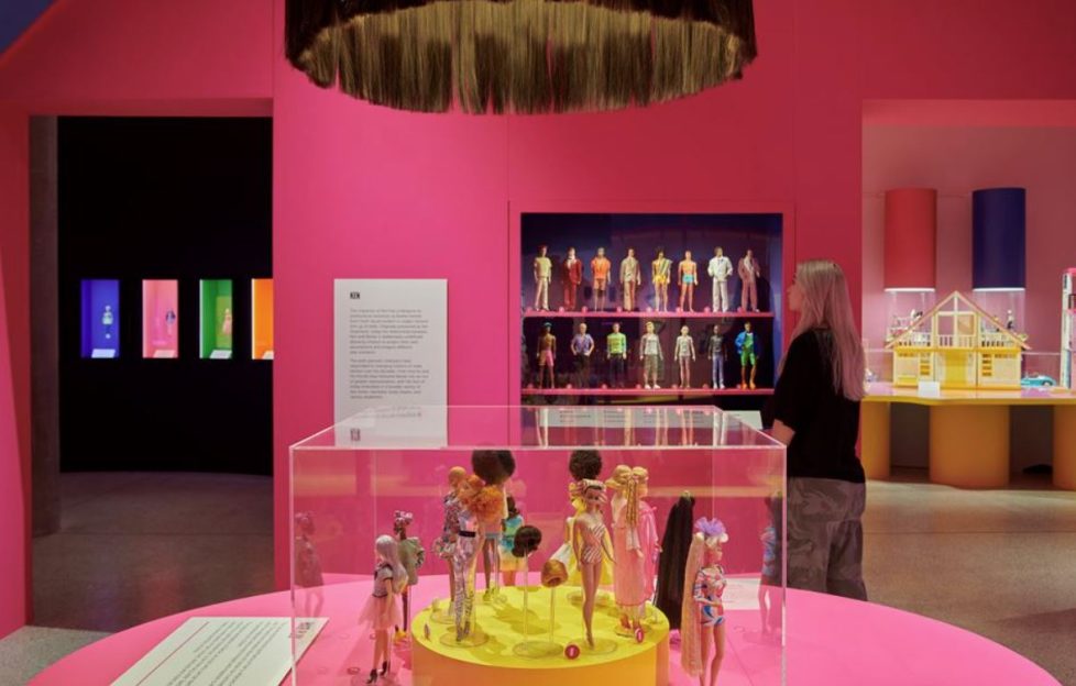 Photo from inside the Barbie exhibition London. There is a chandelier made of barbie hair, every wall is Barbie pink, and there are dolls of all variety in different types of casing spaced out across the room.