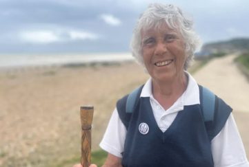 Successful people later in life. Pam Brooks who completed a 90-mile charity walk is seen standing on the beach front holding a walking pole having completed her trek. She is smiling wide.