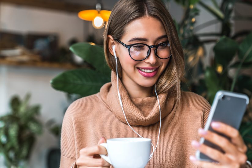 Young smiling woman holding cup of coffee. She's wearing headphones and looking at phone.