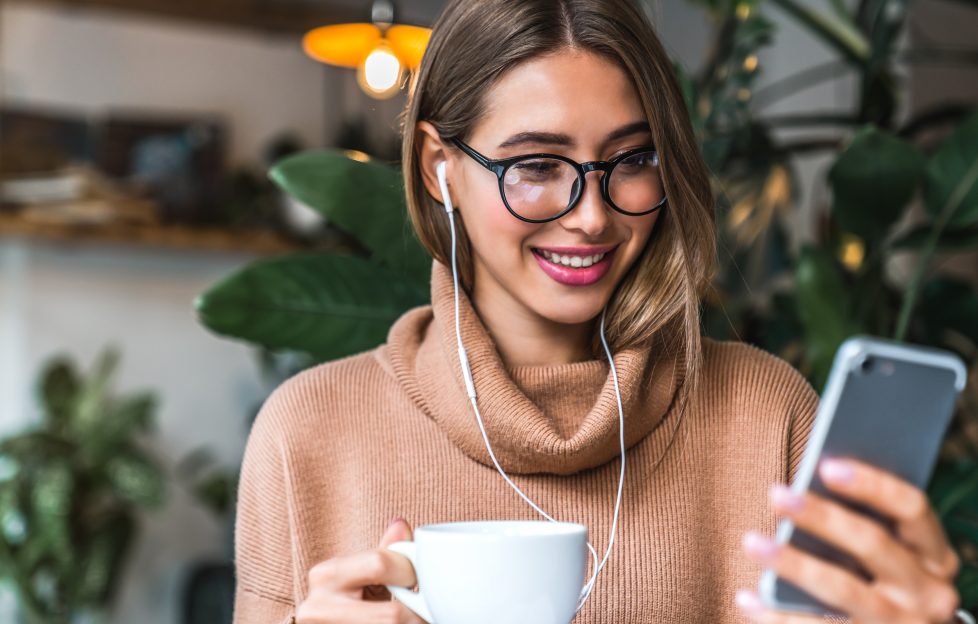 Young smiling woman holding cup of coffee. She's wearing headphones and looking at phone.