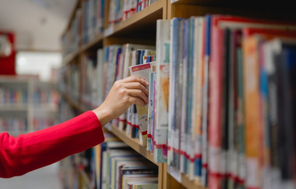 public libraries funding crisis. woman reaches into a library book shelf to pick a book.