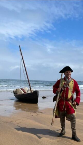 Aaron on a beach, in front of a paddle boat in his redcoat costume with a long rifle prop.