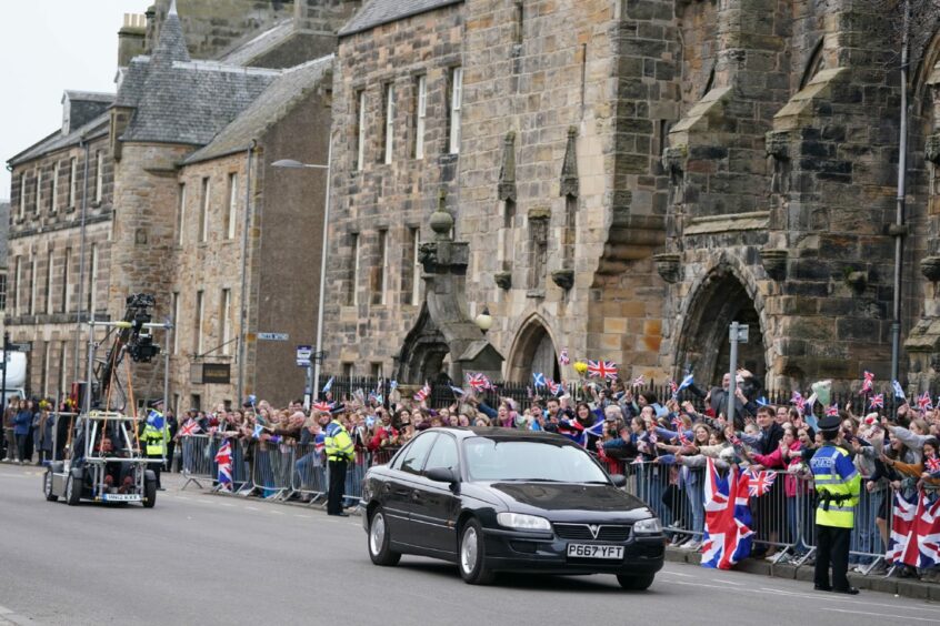 Prince William travelling by car through a St Andrews street past a crowd of background actors as part of a scene in Netflix show The Crown.