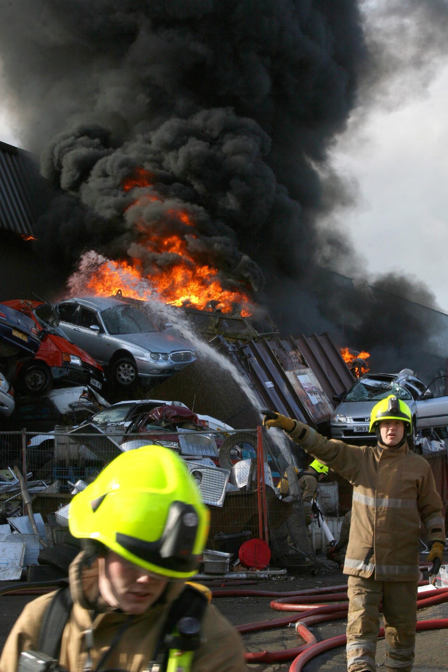 Flames rise into the air behind a stack of cars in the scrapyard. 