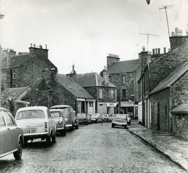 Shops and cars parked on both sides of the cobbled street. Image: DC Thomson.