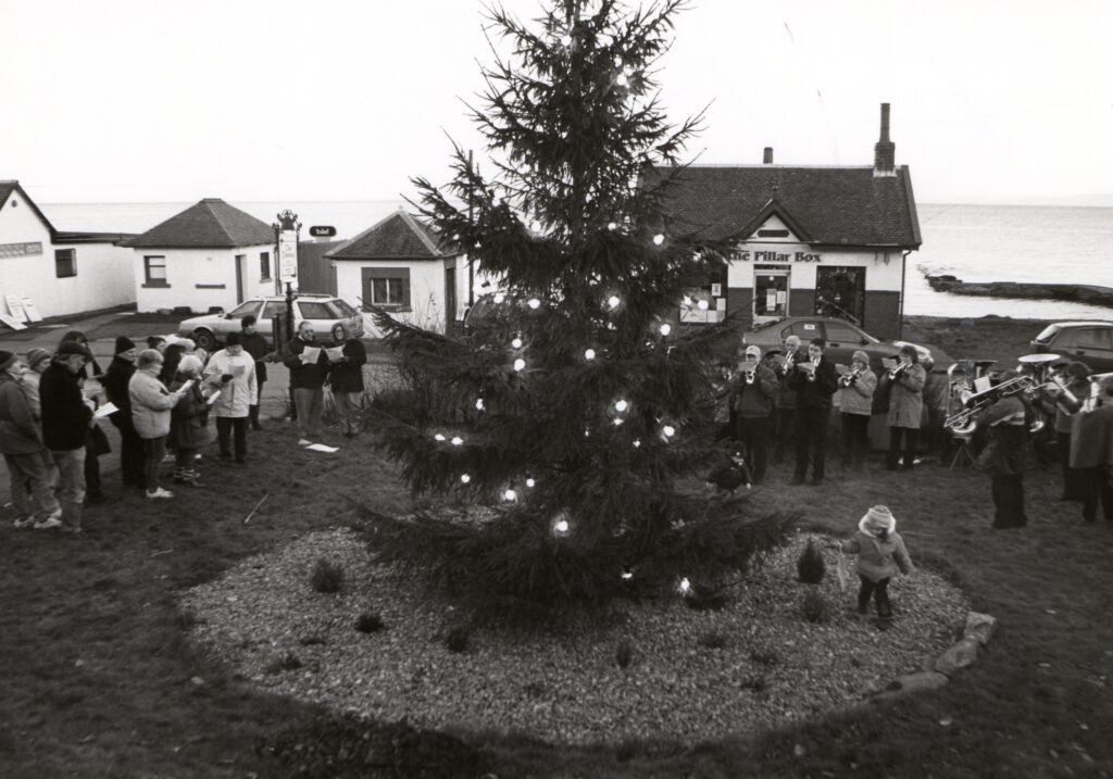 A small group of carol singers enjoy carolling around the Christmas tree in Whiting Bay accompaniment by Arran Brass.