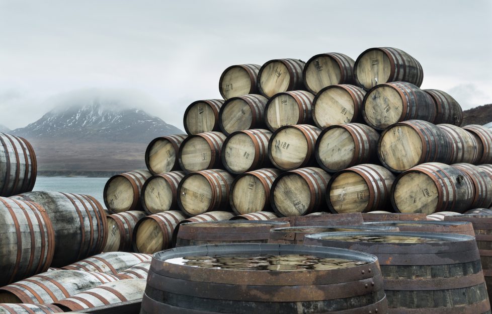 Scotland's Whisky Regions. Image features whisky casks stockpiled outside Bunnahabhain distillery Islay with a snowy mountain across the water as a backdrop. The casks are waiting to be bottled.