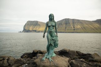 Selkie statue, Mikladalurs Kopakonan, on the Faroe Islands. A sculpture of a naked woman stands on the shoreline holding a seal skin which she is stepping out of.
