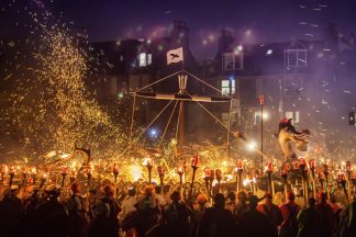 up helly aa included women this year. Image shows a ship in the process of being burned. Embers are flying across a purple, smokey sky. In the foreground procession participants stand holding burning torches as they watch the ship burn.