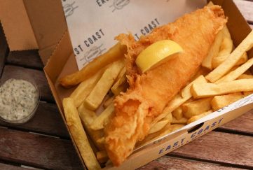 fish and chips from a Scottish fish and chip shop. The food is in a box with a side of tartar sauce on a wooden table.