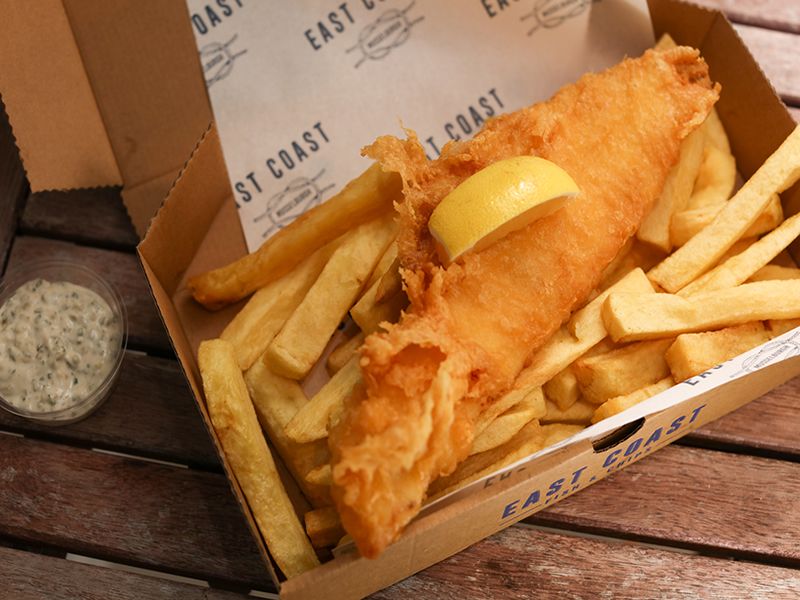 fish and chips from a Scottish fish and chip shop. The food is in a box with a side of tartar sauce on a wooden table.