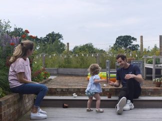 Image of Andy playing out in the garden with his daughter. His wife Kim sits watching them from a small flowerbed wall. 