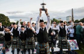 Images shows the Boghall and Bathgate Caledonia Pipe Band winning at the 2023 World Pipe Band Championships. The band are full dress in their tartan and one band member is holding the trophy up high above his head while they all celebrate.