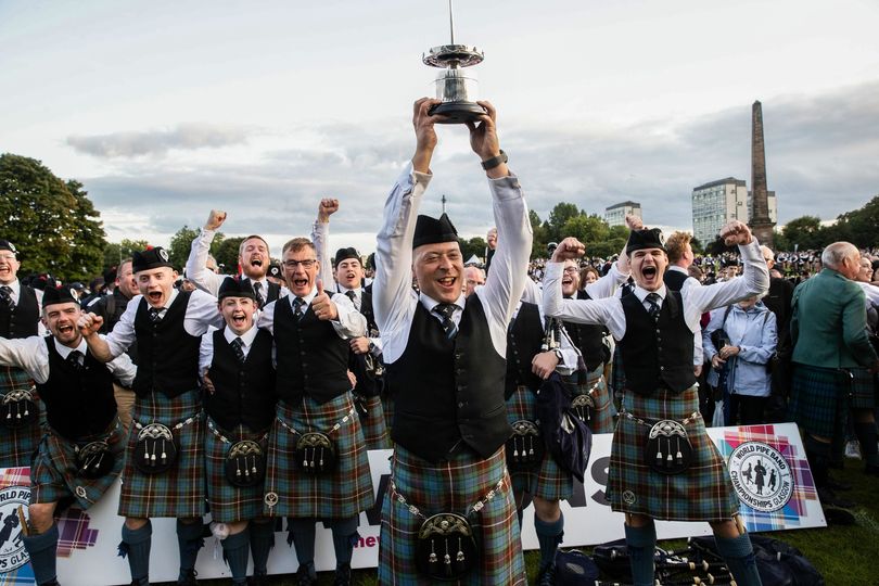 Images shows the Boghall and Bathgate Caledonia Pipe Band winning at the 2023 World Pipe Band Championships. The band are full dress in their tartan and one band member is holding the trophy up high above his head while they all celebrate.