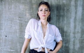 Scottish singer KT Tunstall posing against a concrete wall. She wears a loose white shirt and black trousers