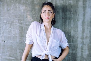 Scottish singer KT Tunstall posing against a concrete wall. She wears a loose white shirt and black trousers