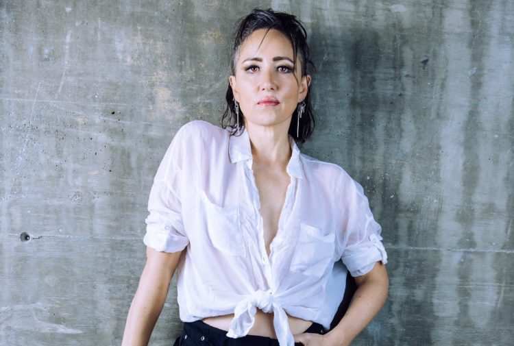 Scottish singer KT Tunstall posing against a concrete wall. She wears a loose white shirt and black trousers