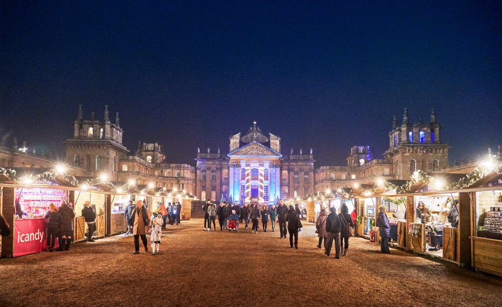 Blenheim Palace courtyard at night with lit wooden cabins and visitors