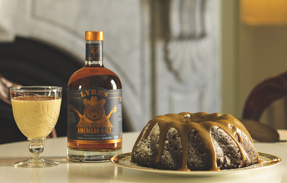 ring shaped fruit cake with caramel sauce, bottle of spirit and glass of eggnog on table