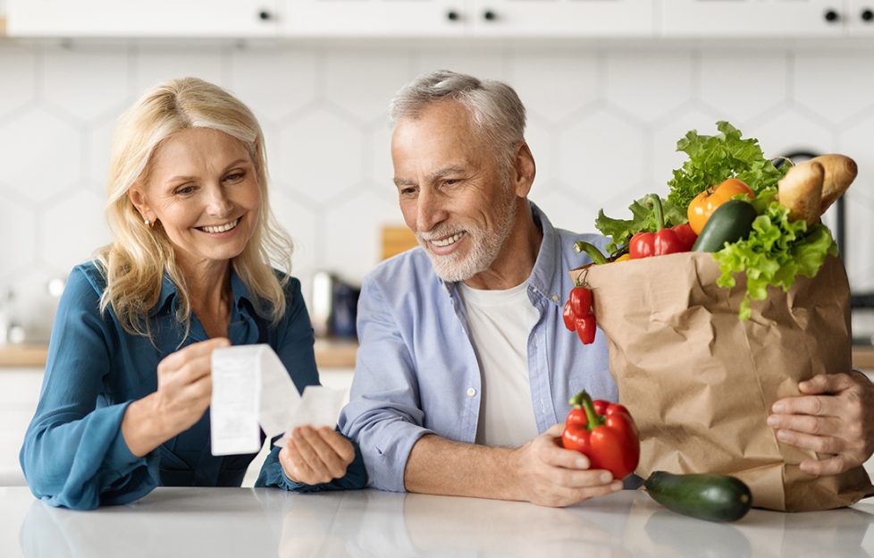 A mature husband and wife looking at grocery bill with fresh veg beside them on their kitchen worktop