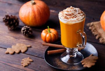 pumpkin spiced latte in glass topped with cream, on stained wooden table with small pumpkins, cinnamon sticks and ginger biscuits