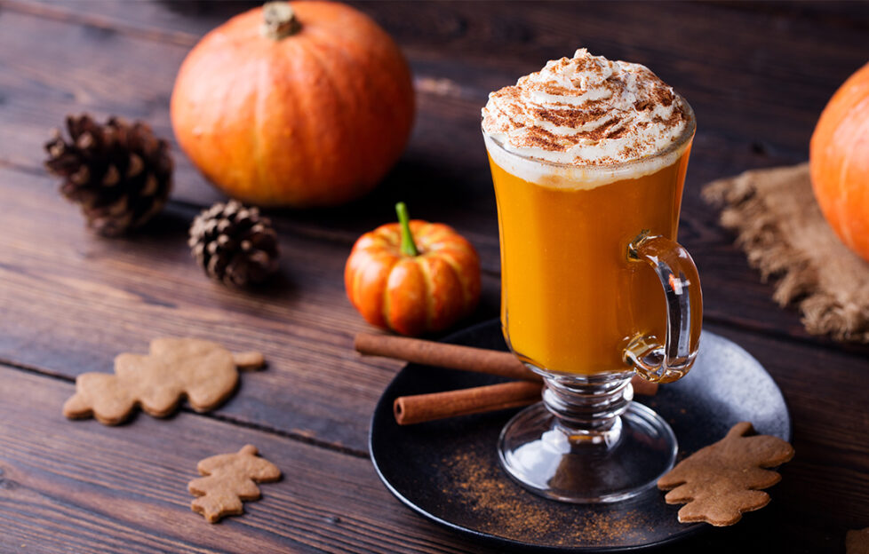 pumpkin spiced latte in glass topped with cream, on stained wooden table with small pumpkins, cinnamon sticks and ginger biscuits