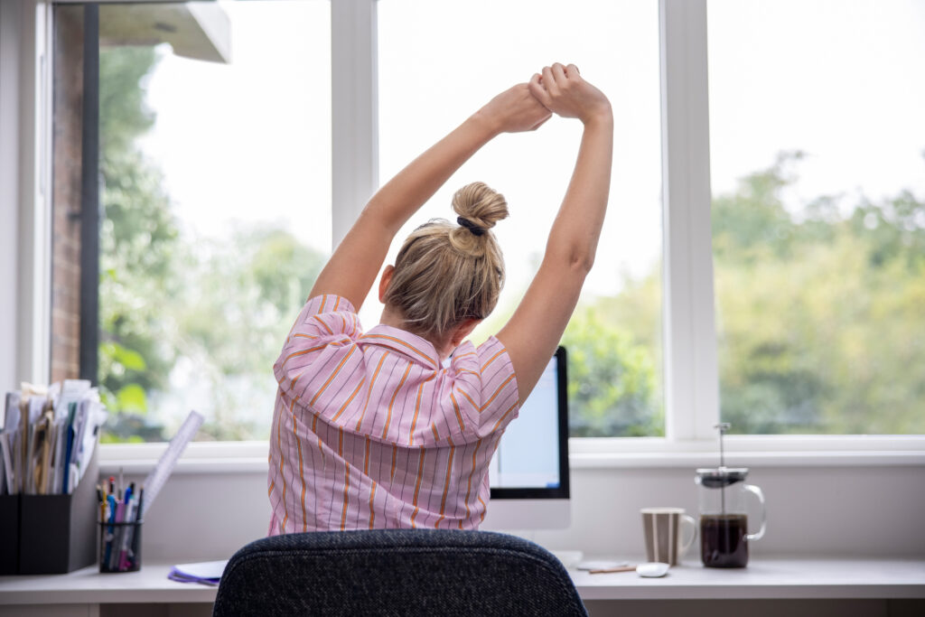 Rear View Of Woman Working From Home On Computer In Home Office Stretching At Desk; 