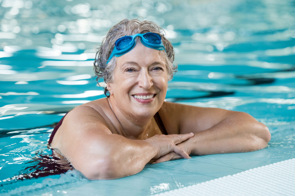 Mature woman wearing swim goggles at swimming pool. Fit active senior woman enjoying retirement in swimming pool and looking at camera. Happy senior healthy old woman enjoying active lifestyle.