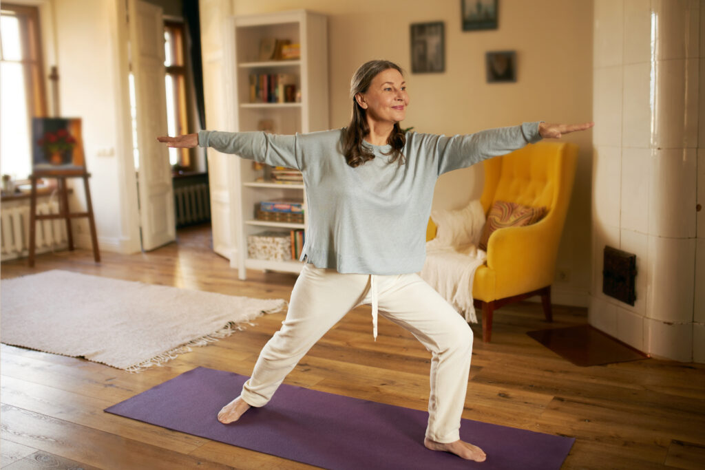 Full length shot of happy energetic mature woman in casual clothes exercising at home because of social distancing, practicing yoga on mat, standing in warrior ii pose. Age, wellness and health; 