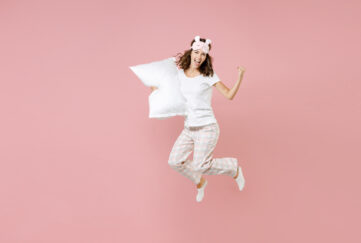 Full length portrait of happy young woman in white pajamas home wear jumping hold pillow doing winner gesture rest at home isolated on pastel pink background studio. Relax good mood lifestyle concept;