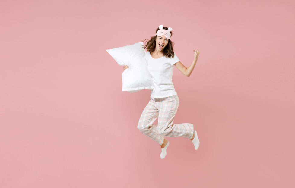 Full length portrait of happy young woman in white pajamas home wear jumping hold pillow doing winner gesture rest at home isolated on pastel pink background studio. Relax good mood lifestyle concept;