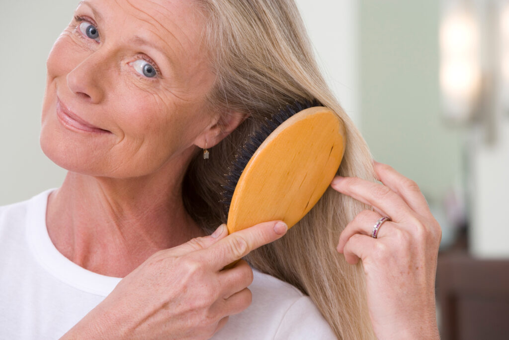 Close-up portrait of a smiling senior woman brushing long straight hair with a hairbrush in the bathroom.; 