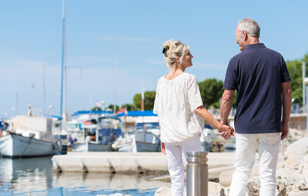 Mature couple enjoying a day at the coast walking away from the camera hand in hand past a small boat harbour;