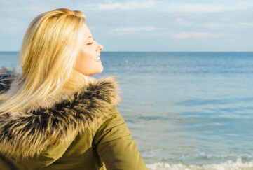 Leisure, spending free time outside, healthy walks concept. Woman wearing warm jacket relaxing on beach near sea, cold sunny day, back view;
