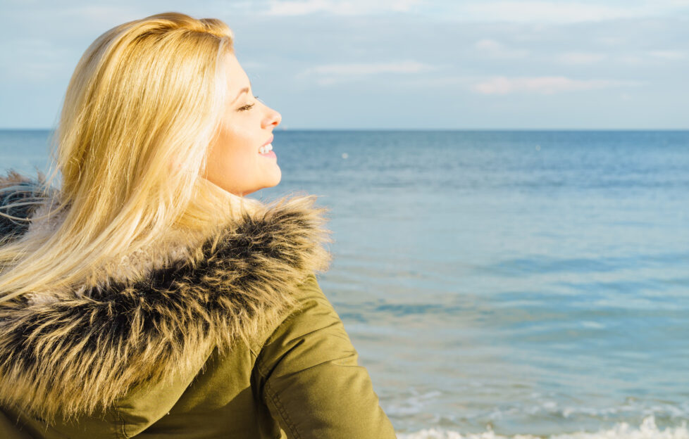 Leisure, spending free time outside, healthy walks concept. Woman wearing warm jacket relaxing on beach near sea, cold sunny day, back view;