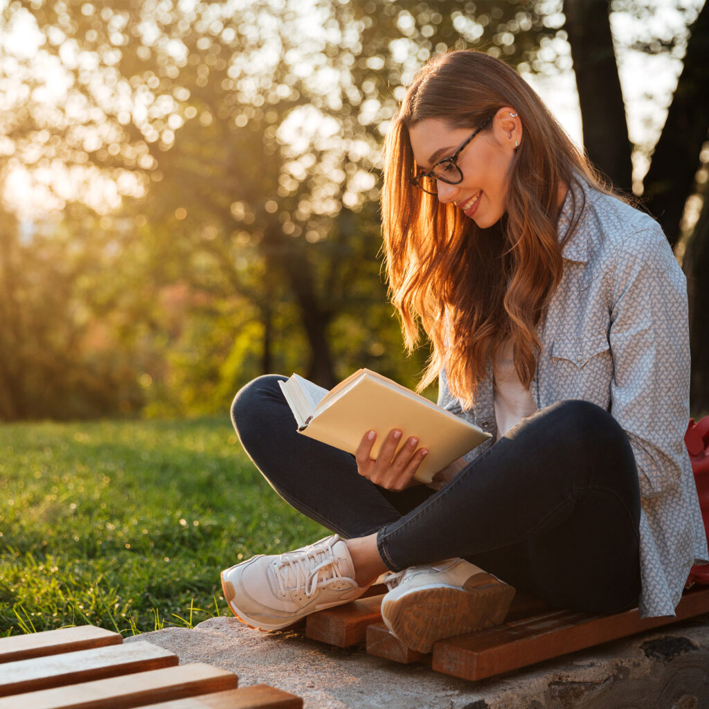 Side view of brunette woman in eyeglasses sitting on bench and reading book in park