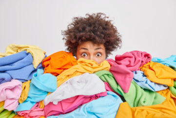 Shocked woman with curly Afro hair stares bugged eyes drowned in huge pile of colorful clothing cleans out closet selects clothes for donation or recycling white background. Housework concept;