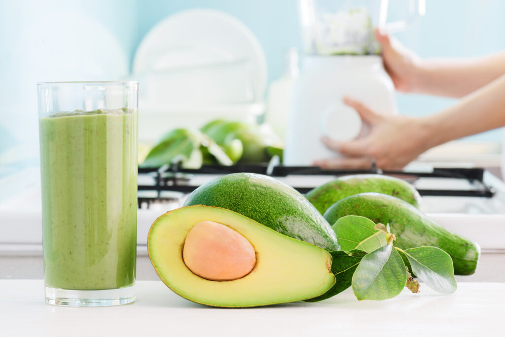 Fresh avocado smoothie and ripe green avocados on kitchen table. Hands of woman using blender are visible in background. Healthy eco food and natural drink.; 
