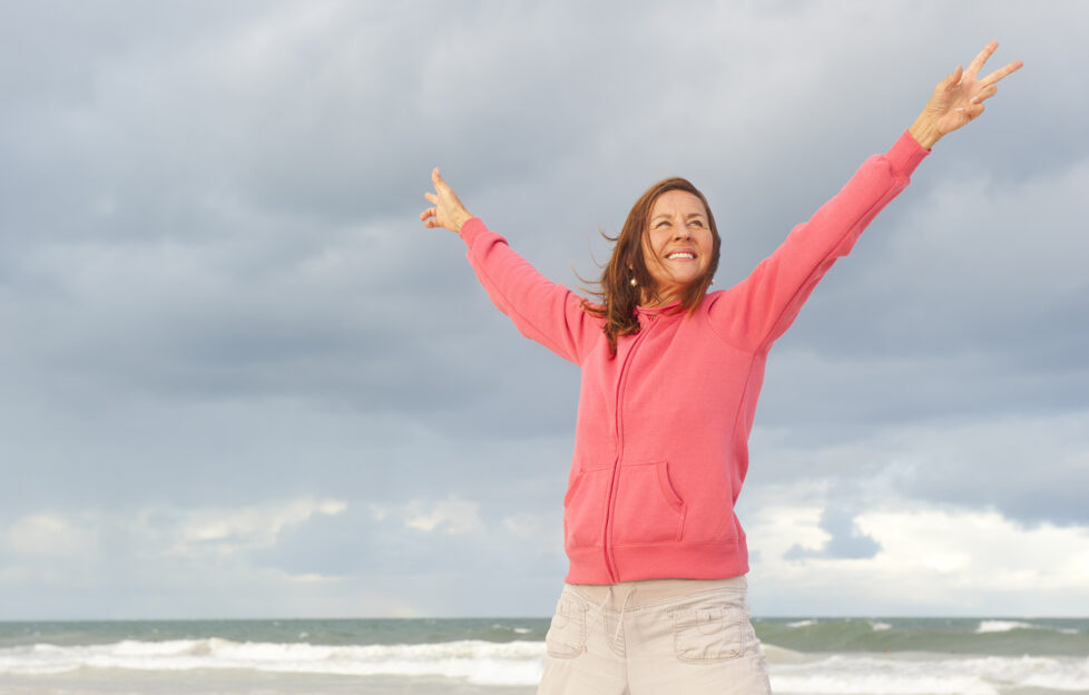 Portrait of positive and happy mature woman at the beach, wearing pink sweater, isolated with ocean and storm clouds