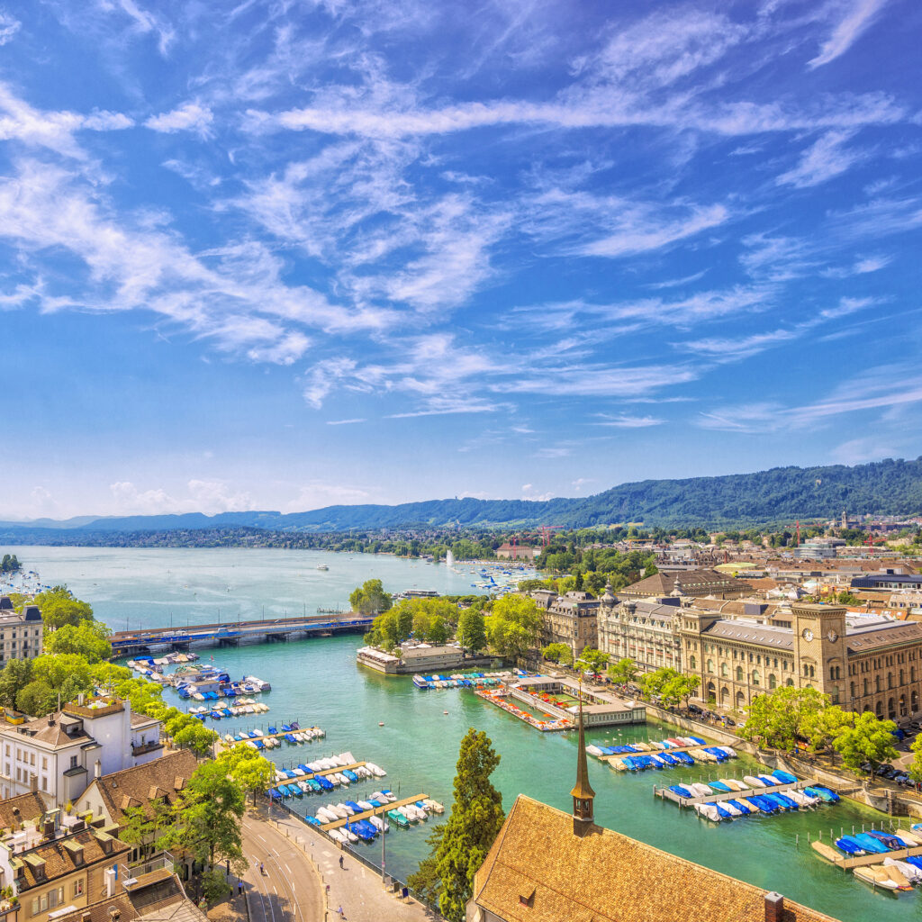 Elevated view over the old town of Zurich with the Stadthausquai and traditional public Limmat river bath (Frauenbad), the lake Zurich and the Swiss alps in the distance on a beautiful summer day.