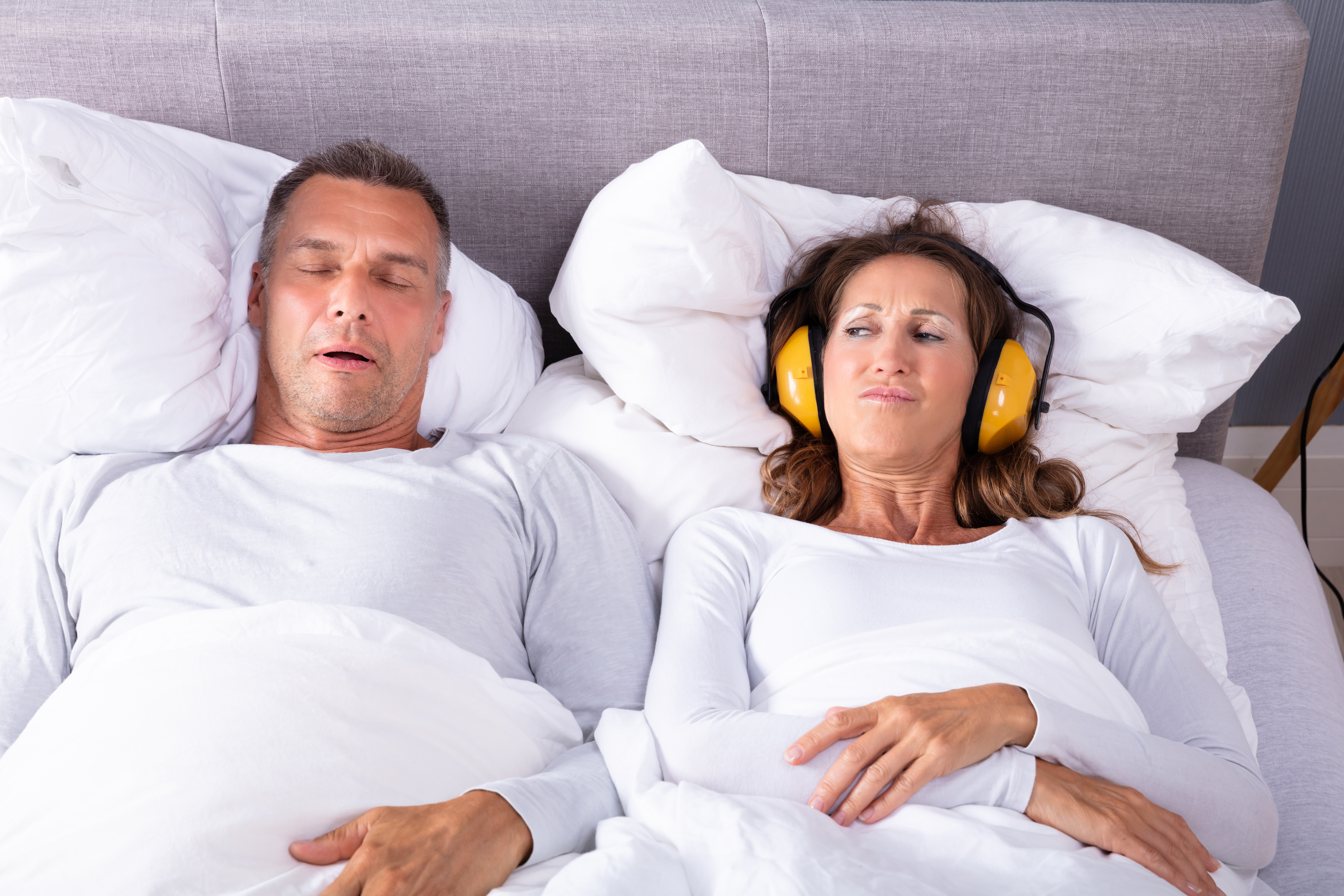 Mature Woman Covering Her Ears With Headphone While Man Snoring In Bed